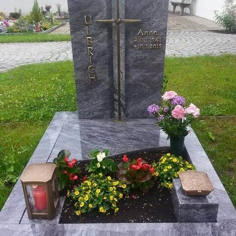 Dunkles Grabmal mit erhaben herausgearbeitetem Kreuz, davor ein bunt bepflanztes Beet.