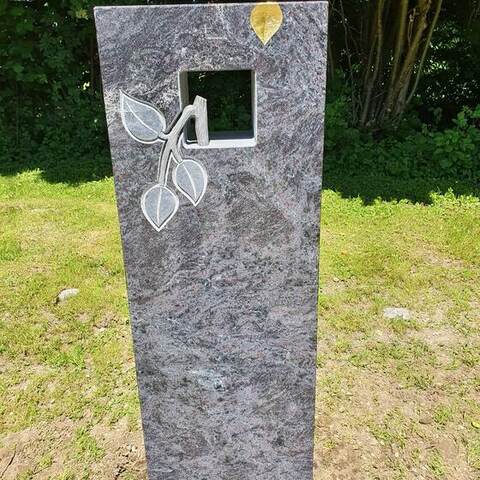 Graue Stele mit einem quadratischen Durchbruch und einem aufgesetzten Blattmotiv aus Metall.