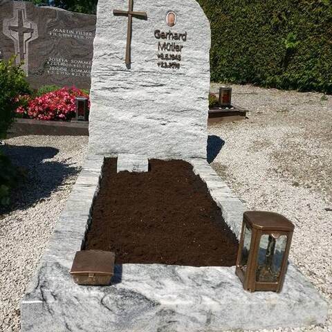 Heller Stein mit bruchrauen Kanten, eingehauenem Kreuz und Schrift auf einer Grabanlage mit Einfassung.