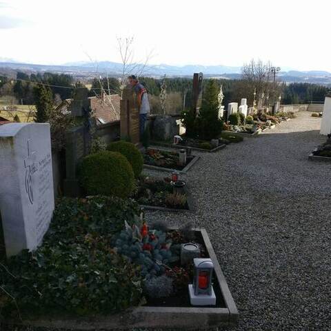 Kiesweg zwischen Grabreihen auf einem Friedhof, im Hintergrund die Berge des Allgäus.