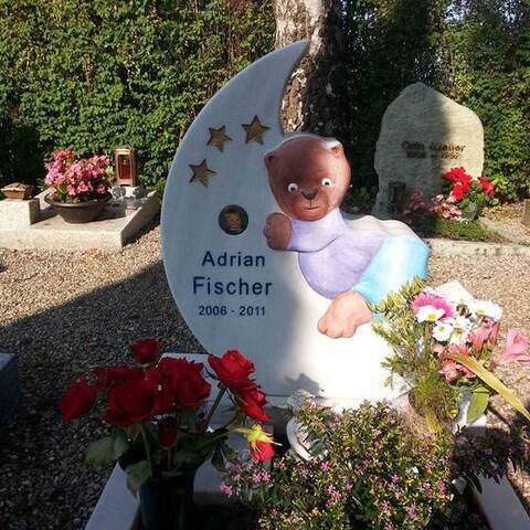 Helles, mondförmiges Kindergrabmal mit aufgesetzter Figur und vergoldeten Sternen, umgeben von Blumen.