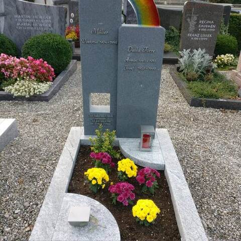 Graue Stele mit einem aus farbigem Glas eingelegten Regenbogen, davor ein bunt bepflanztes Beet.