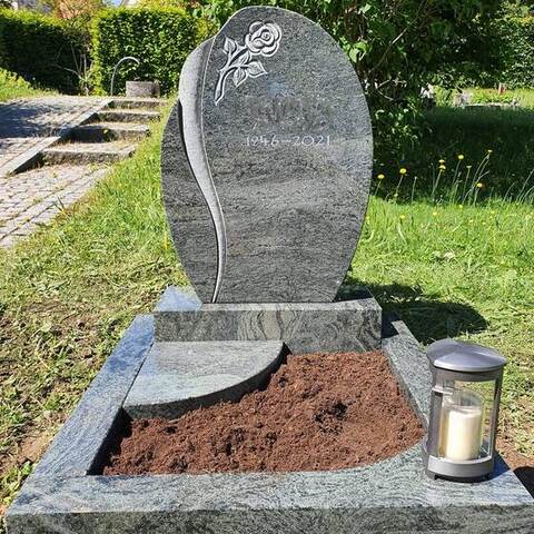 Rundbogiges graues Grabmal mit geschliffener Rose, davor ein frisch angelegtes Beet und eine Laterne.