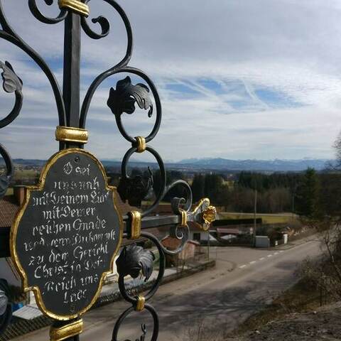 Schmiedeeisernes Grabkreuz mit vergoldeter Schrifttafel, dahinter der Blick ins Voralpenland.