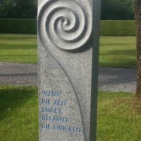 Ausschnitt einer hellen Granitstele mit Spirale und von Hand geschlagener, blau gefasster Inschrift.