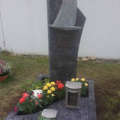 Frei geformte, geschwungene Skulptur aus dunklem Stein auf einer Grabanlage.