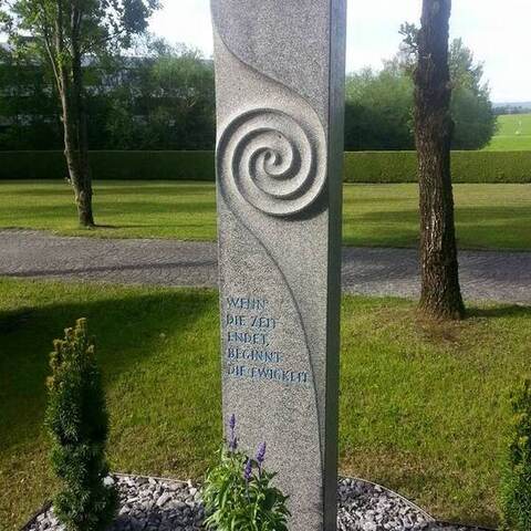 Hohe helle Granitstele mit eingearbeiteter Spirale und blau gefasster Inschrift, davor eine Bepflanzung im Kiesbett.