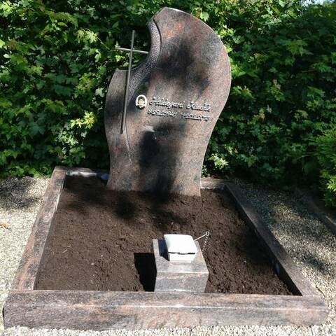 Geschwungenes Grabmal aus rotbraunem Granit mit eingelassenem Medaillon und Kreuz, davor ein frisch angelegtes Beet.