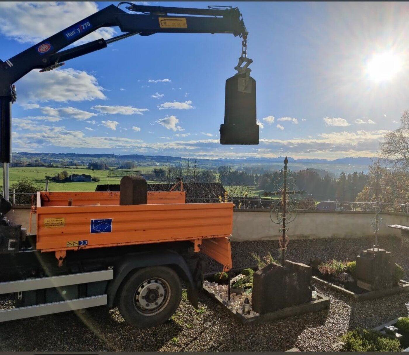 Orangefarbenes Kranfahrzeug hebt einen Grabstein über die Friedhofsmauer, im Hintergrund das Allgäuer Voralpenland.