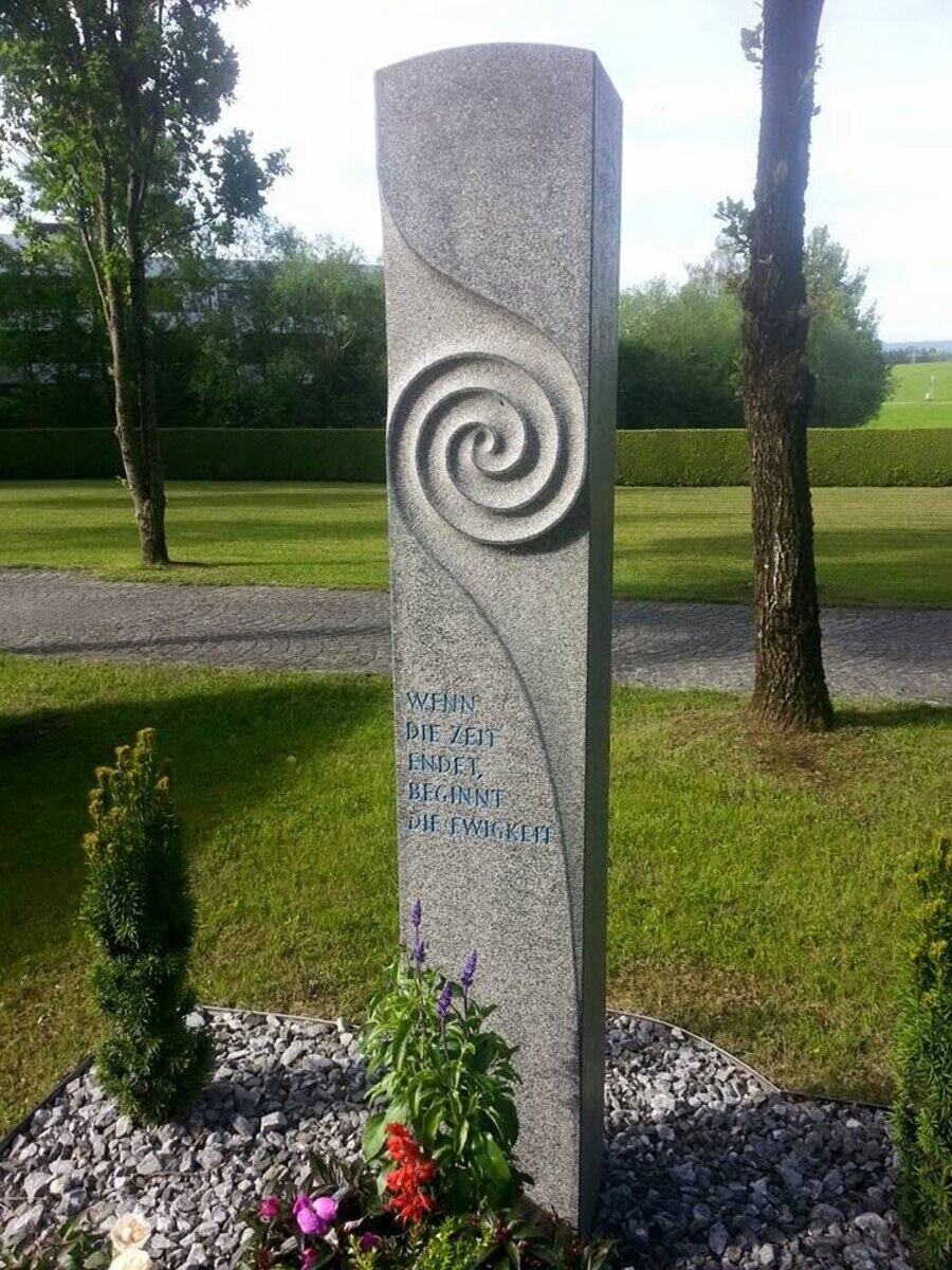 Hohe helle Granitstele mit eingearbeiteter Spirale und blau gefasster Inschrift, davor eine Bepflanzung im Kiesbett.