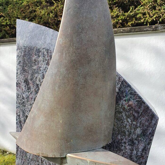 Segelförmige Bronzeskulptur auf einem hellen Steinsockel vor einer dunklen Stele.