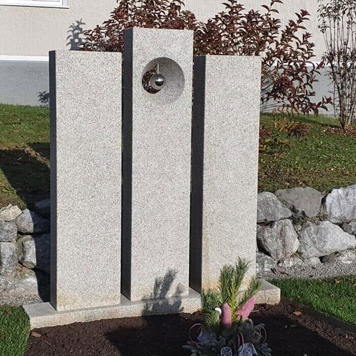 Drei dunkle Stelen unterschiedlicher Höhe mit einer Kugel im Durchbruch, in einem Garten aufgestellt.