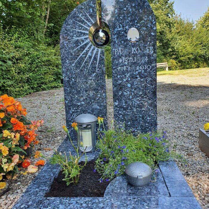 Zweiteiliges Grabmal aus blau-schwarzem Granit mit gestockter Strahlengravur und einem goldfarbenen Ring.
